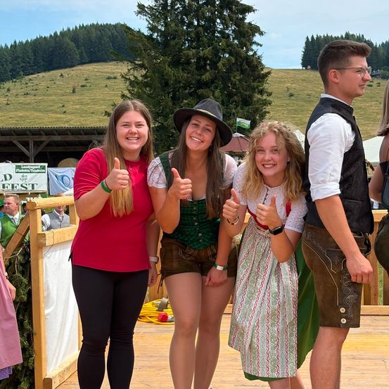 Vier junge Leute, zwei Frauen und zwei Männer, stehen zusammen auf einer Holzplattform, lächeln und geben Daumen hoch. Sie sind in traditioneller deutscher Kleidung gekleidet. Hinter ihnen befindet sich ein Holzzaun, ein Baum und ein grasbewachsener Hügel.