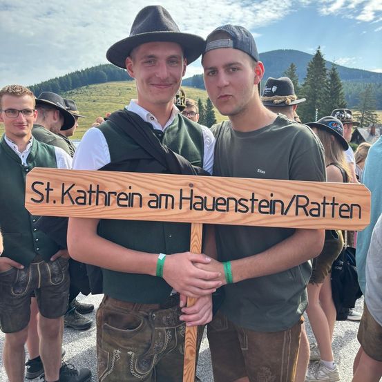 Zwei Männer in traditioneller bayerischer Kleidung stehen hinter einem Holzschild, das St. Kathrein am Hauenstein/Ratten besagt. Sie lächeln und posieren für ein Foto.
