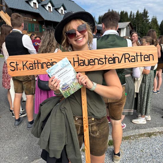 Eine Frau hält ein Holzschild mit den Namen St. Kathrein und Hauenstein/Ratten und lächelt für ein Foto in einer festlichen Umgebung mit traditioneller Kleidung im Hintergrund.