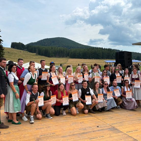 Eine Gruppe von Menschen in traditioneller Kleidung, die Urkunden halten, posiert für ein Foto auf einer Holzbühne mit einem Gebirgshintergrund.