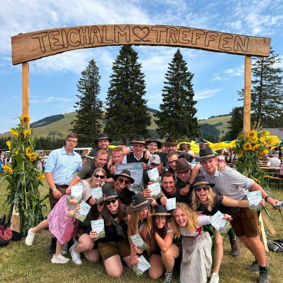 Eine Gruppe von Menschen posiert für ein Foto vor einem Holzbogen, der Teichalm2treffen steht. Sie halten Urkunden und tragen Hüte. Im Hintergrund sind Sonnenblumen und Bäume zu sehen.