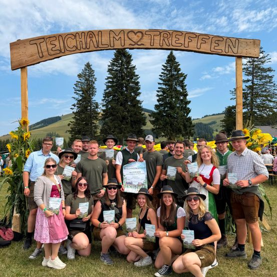Eine Gruppe von Menschen in traditioneller Kleidung posiert für ein Foto unter einem Holzbogen mit den Worten Teichalm Treffen. Sie halten Urkunden in der Hand und stehen vor einer malerischen Kulisse.