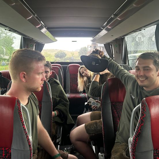 Eine Gruppe von Menschen in einem Bus, ein Mann hält einen Hut über den Kopf einer Frau und lächelt. Der Bus fährt auf einer Straße, draußen sind Bäume und Gebäude zu sehen.
