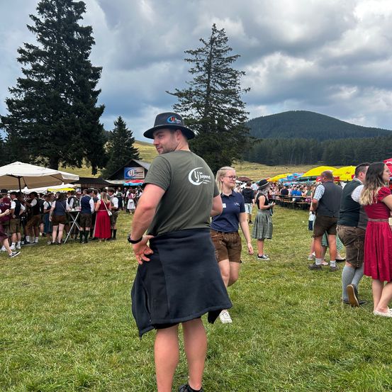 Ein Mann in einem Kilt steht auf einem Grasfeld bei einem Festival mit Menschen in traditioneller bayerischer Kleidung, Bergen im Hintergrund und bunten Regenschirmen.