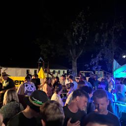 Eine nächtliche Zusammenkunft mit Menschen in einem Park, einige tragen Hüte und werden von blauen Lichtern beleuchtet. Bäume und ein gelbes Banner sind im Hintergrund zu sehen.