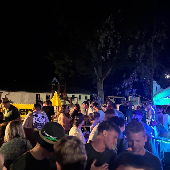 Eine nächtliche Zusammenkunft mit Menschen in einem Park, einige tragen Hüte und werden von blauen Lichtern beleuchtet. Bäume und ein gelbes Banner sind im Hintergrund zu sehen.