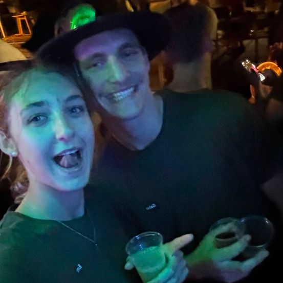 Ein Mann und eine Frau lächeln und posieren für ein Foto auf einer Party, mit Getränken in den Händen.
