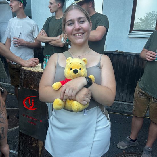 Eine Frau in einem weißen Rock und einem grauen Tanktop hält einen gelben Teddybären, lächelt und steht vor einer Gruppe von Menschen.