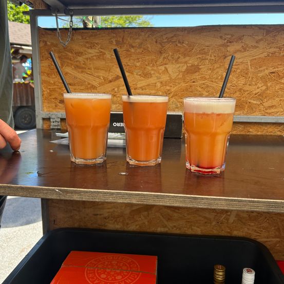 Drei Gläser mit orangefarbenen Getränken und schwarzen Strohhalmen stehen auf einem Holztisch. Der Tisch befindet sich in einem Food Truck mit einer Holzwand. Unten links steht ein rotes Kästchen.