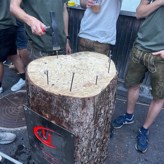 Ein Mann schlägt Nägel in einen Baumstumpf, während eine Menge zuschaut. Der Baumstumpf hat ein Schild mit einem roten Logo.