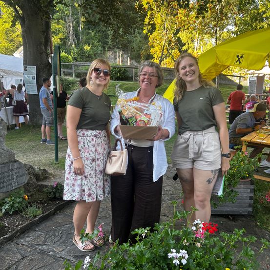 Drei Frauen stehen in einem Garten, eine hält einen Geschenkkorb. Sie lächeln und scheinen für ein Foto zu posieren. Hinter ihnen sitzen Leute an einem Picknicktisch unter einem gelben Schirm.