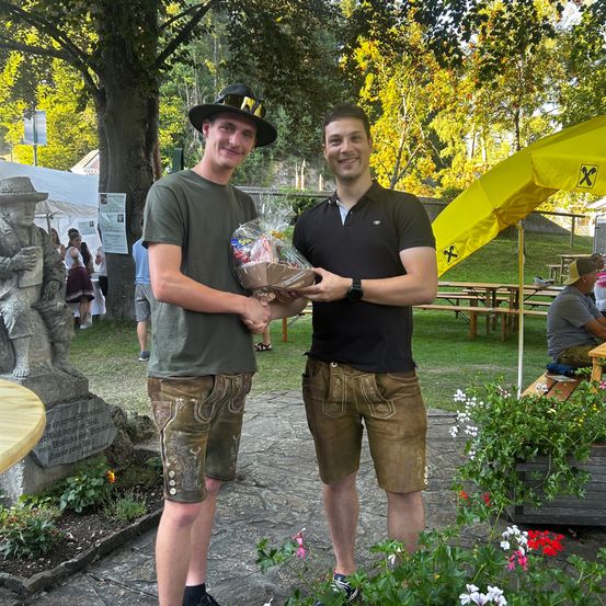 Zwei Männer in traditioneller bayerischer Kleidung stehen in einem Park, einer hält einen Geschenkkorb und der andere lächelt. Hinter ihnen steht eine Statue, ein gelber Regenschirm und ein Picknickbereich mit Tischen und Bänken.