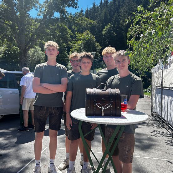 Fünf junge Männer stehen um einen kleinen Tisch mit einer Holzkiste herum, tragen passende grüne Hemden und Shorts. Sie befinden sich in einer Außenumgebung mit Bäumen und einem weißen Zelt im Hintergrund.