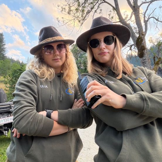 Zwei Frauen in passenden grünen Jacken und Hüten stehen in einem Wald, eine hält ein Funkgerät. Sie lächeln und scheinen für ein Foto zu posieren.