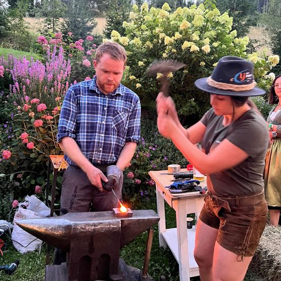 Ein Mann und eine Frau arbeiten in einem Garten an einem Metall Amboss, umgeben von Blumen und einem Holztisch.