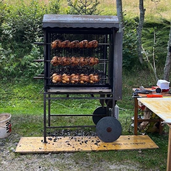 Ein großer Outdoor-Grill mit mehreren Hähnchen, die auf Metallstangen gebraten werden, auf einer Holzplattform aufgestellt, mit einem Eimer in der Nähe.
