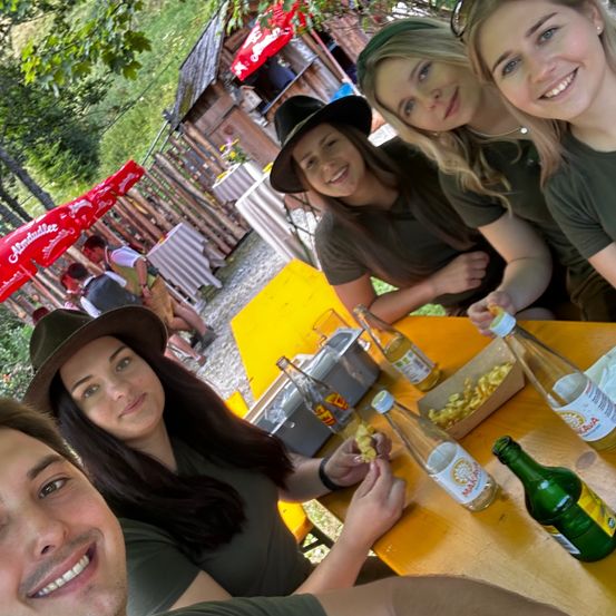 Vier Personen lächeln und posieren für ein Foto in einem Restaurant im Freien. Ein Tisch mit Essen und Getränken ist im Vordergrund zu sehen. Dahinter sind ein Holzhaus und ein roter Regenschirm zu erkennen.