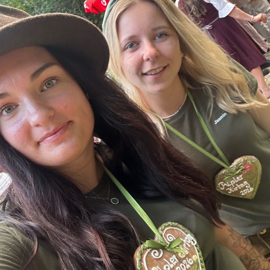Zwei Frauen lächeln und posieren für ein Selfie. Eine hat einen herzförmigen Anhänger mit dem Text 'Baptist 2016'. Die andere hat ein grünes Namensschild mit dem Namen 'Jasmin'. Sie tragen grüne Hemden. Hinter ihnen steht eine weitere Person.
