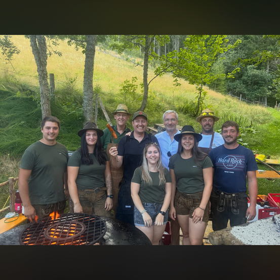 Eine Gruppe von Menschen in traditioneller Kleidung steht vor einem Feuerkorb und lächelt für ein Foto. Im Hintergrund befinden sich eine grasbewachsene Anhöhe und Bäume.