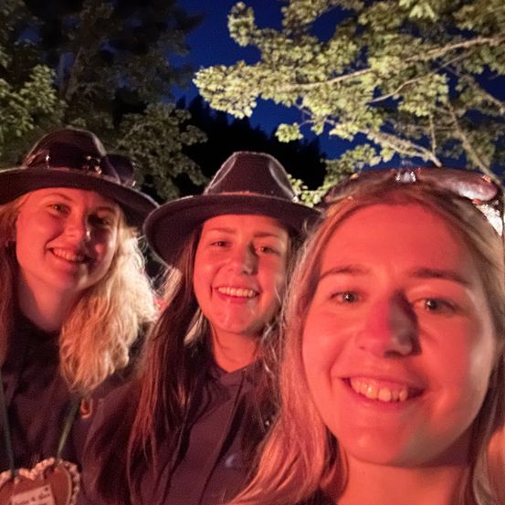 Drei Frauen in passenden Hüten lächeln und posieren nachts für ein Foto mit Bäumen im Hintergrund.