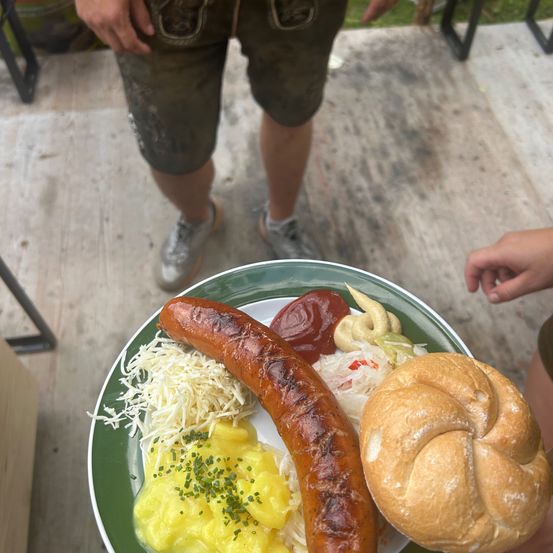 Ein Teller mit einer Wurst, Kartoffelpüree, Sauerkraut und einem Brezel. Der Teller steht auf einer Holzoberfläche.
