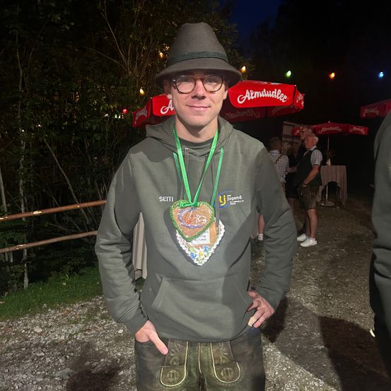 Ein Mann mit Hut und Brille steht nachts im Freien, trägt eine grüne Kapuzenjacke mit einem herzförmigen Abzeichen auf der Vorderseite. Hinter ihm stehen mehrere Personen, und einige Regenschirme sind auf dem Boden platziert.