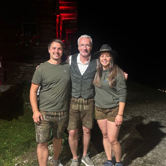 Drei Menschen stehen nachts lächelnd zusammen. Der Mann auf der linken Seite trägt ein grünes Shirt und Shorts mit traditionellem Design. Der Mann in der Mitte trägt eine Weste und eine Brille. Die Frau auf der rechten Seite trägt einen Hut und eine grüne Kapuzenjacke. Hinter ihnen steht ein Gebäude mit roten Lichtern.