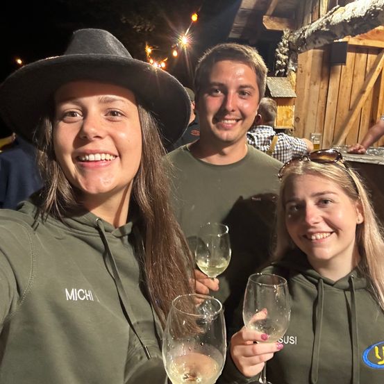 Drei Menschen lächeln und halten Weingläser bei einer Party. Die Frau auf der linken Seite trägt einen Hut und eine grüne Kapuzenjacke mit 'MICHI' darauf. Der Mann in der Mitte trägt ein grünes T-Shirt. Die Frau auf der rechten Seite trägt eine grüne Kapuzenjacke mit 'SUS' darauf. Hinter ihnen steht eine Holzstruktur mit einem Dach.