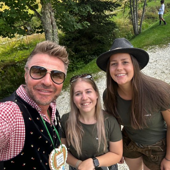 Drei Personen, zwei Frauen und ein Mann, lächeln und posieren für ein Foto auf einem Kiesweg mit Bäumen und einem Grasbereich im Hintergrund.