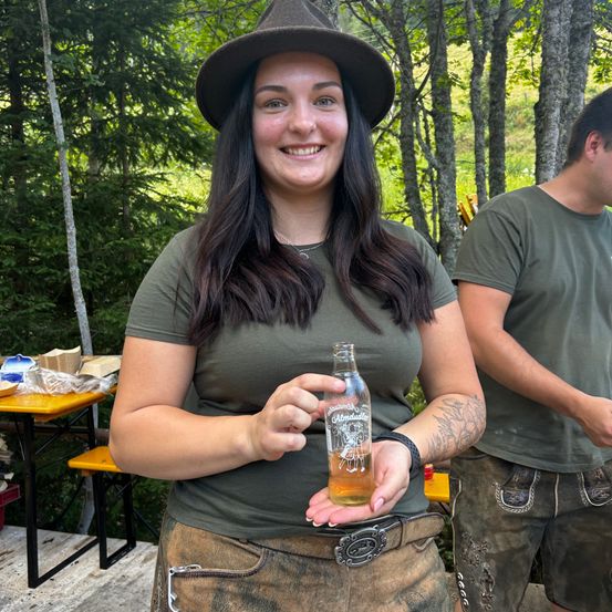 Eine Frau in einem grünen Shirt und Hut hält eine kleine Flasche Bier in einer Waldumgebung, mit einer weiteren Person in traditioneller Kleidung im Hintergrund.