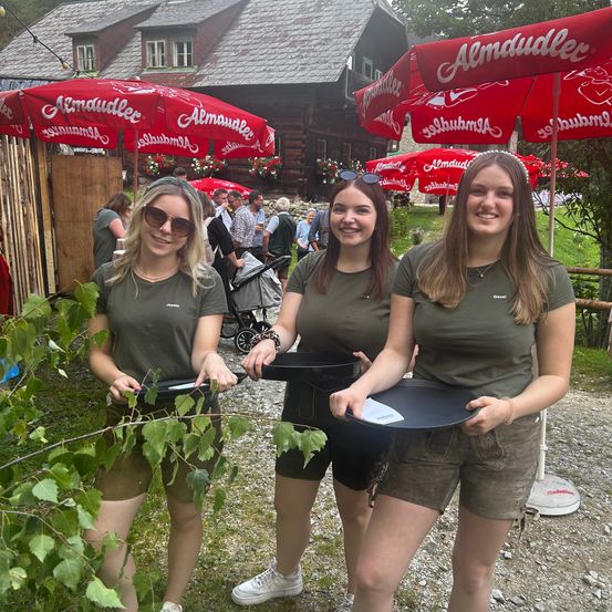 Drei Frauen in grünen Shirts und Shorts posieren mit einem Tablett und einer Pflanze vor einem Gebäude mit roten Sonnenschirmen.