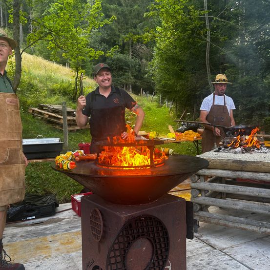 Drei Männer in Schürzen kochen Essen über offenem Feuer in einer Waldumgebung, wobei einer einen Spieß hält.
