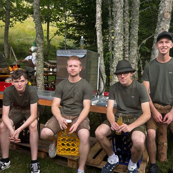 Vier junge Männer in grünen Shirts und Shorts sitzen auf Holzpaletten in einem Wald und halten Gläser mit Bier. Hinter ihnen kocht ein Mann am Feuer.