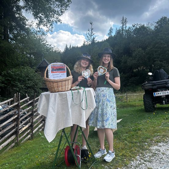Zwei Frauen stehen neben einem Tisch mit einem Korb und herzförmigen Keksen und lächeln in einem Grasbereich mit einem Holzzaun und einem Fahrzeug im Hintergrund.