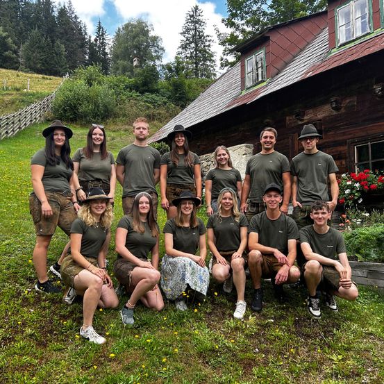 Eine Gruppe von Menschen in passenden grünen Shirts und Shorts, einige mit Hüten, posiert für ein Foto vor einem rustikalen Haus mit rotem Dach. Der Hintergrund zeigt ein Grasfeld mit Blumen und Bäumen.