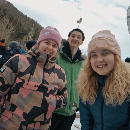 Drei junge Menschen stehen lächelnd für ein Foto in einer schneebedeckten Umgebung. Das Mädchen links trägt eine Camouflage-Jacke, die mittlere Person eine grüne Jacke und das Mädchen rechts eine pinke Mütze.