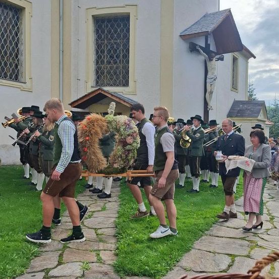 Eine Gruppe von Menschen in traditioneller bayerischer Kleidung, die eine Blumenarrangement auf einem hölzernen Tragbahre tragen, mit einem Blasorchester und Zuschauern im Hintergrund.