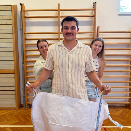 Ein Mann hält einen großen weißen Sack, während zwei Frauen hinter ihm stehen und alle lächeln in einem Raum mit Holzwänden und einem Holzboden.