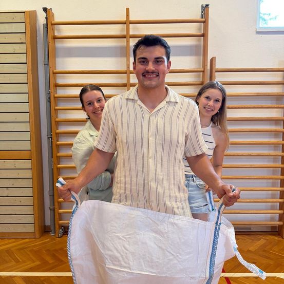 Ein Mann hält einen großen weißen Sack, während zwei Frauen hinter ihm stehen und alle lächeln in einem Raum mit Holzwänden und einem Holzboden.