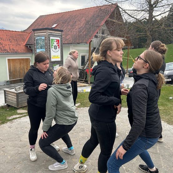 Eine Gruppe von Frauen in Jacken und Turnschuhen versammelt sich vor einem Haus mit rotem Dach, eine Frau beugt sich nach unten, während eine andere nach oben schaut.