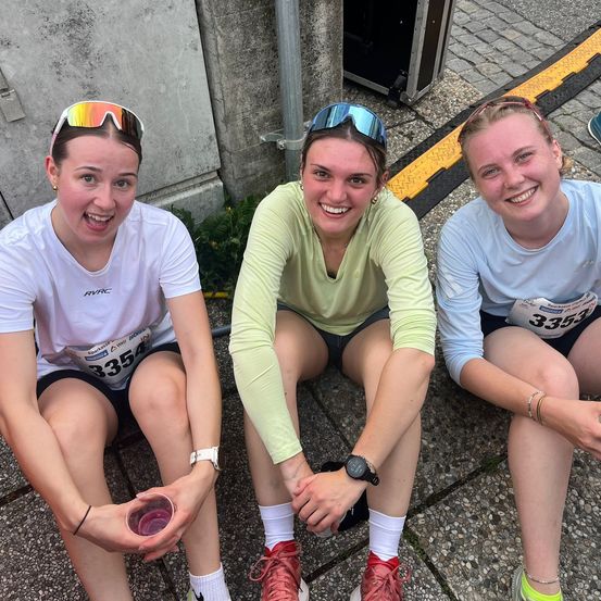 Drei Frauen in Laufausrüstung, einschließlich Sonnenbrillen und Uhren, sitzen auf einer Steinoberfläche und lächeln für ein Foto. Sie haben Marathonnummern auf ihren Shirts.