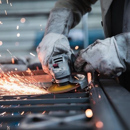 Eine Person mit Handschuhen benutzt eine Schleifmaschine, um Metall zu schneiden, wobei Funken in einer Werkstatt herumfliegen.