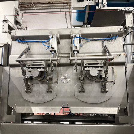 Eine Nahaufnahme einer industriellen Edelstahlmaschine mit mehreren mechanischen Teilen und blauen Kabeln. Die Maschine wird wahrscheinlich für die Herstellung oder Verpackung in einer Fabrik verwendet.