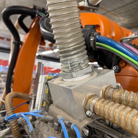 Eine Nahaufnahme einer industriellen Maschine zeigt einen zylindrischen Bauteil, der an einem quadratischen Metallblock befestigt ist, mit mehreren Kabeln und Schläuchen.