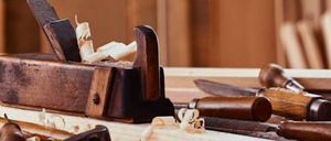 Ein Holzarbeitstisch beherbergt verschiedene Werkzeuge, darunter ein Meißel, eine Hobel und ein Messer. Die Werkzeuge sind von Holzspänen umgeben.