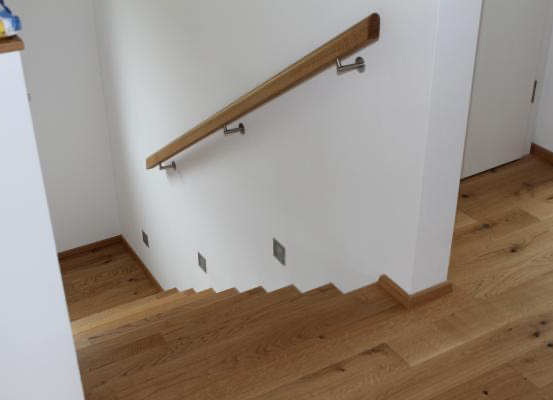 Innenansicht eines Hauses mit Holztreppe und einer weißen Wand mit einem Holzgeländer und Metallgriffen.