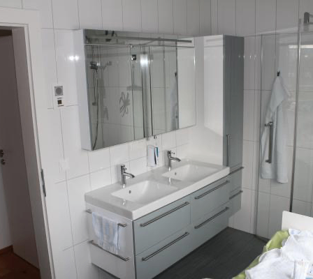 Ein modernes Badezimmer mit einem Doppelwaschbecken, grauen Schränken und einer Glasdusche. Ein Handtuch hängt an der Wand, und es gibt eine Steckdose an der Wand.