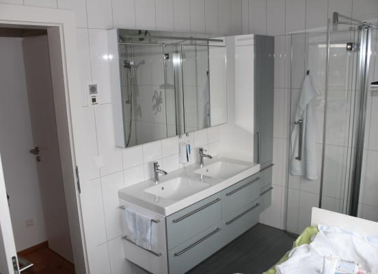 Ein modernes Badezimmer mit einem Doppelwaschbecken, grauen Schränken und einer Glasdusche. Ein Handtuch hängt an der Wand, und es gibt eine Steckdose an der Wand.