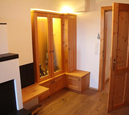 Ein Raum mit Holzboden und einem Holzschrank mit Glastüren. Der Schrank hat eine Lampe im Inneren. Es gibt eine Holzbank und einen Kamin. Eine offene Tür führt in einen anderen Raum.