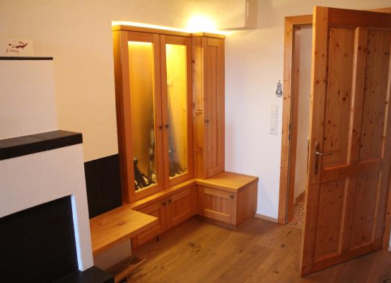 Ein Raum mit Holzboden und einem Holzschrank mit Glastüren. Der Schrank hat eine Lampe im Inneren. Es gibt eine Holzbank und einen Kamin. Eine offene Tür führt in einen anderen Raum.